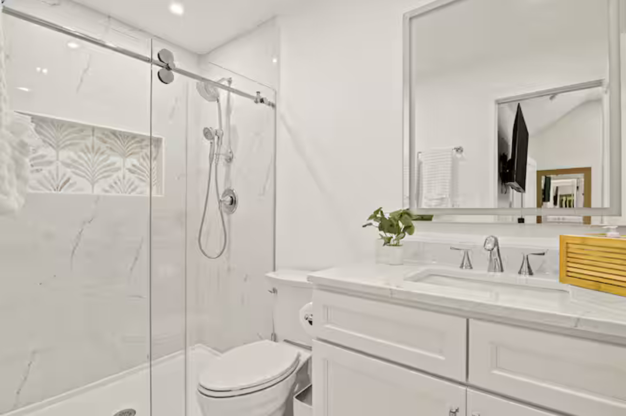 Bright white bathroom with glass shower, vanity sink, mirror, and small plant accents