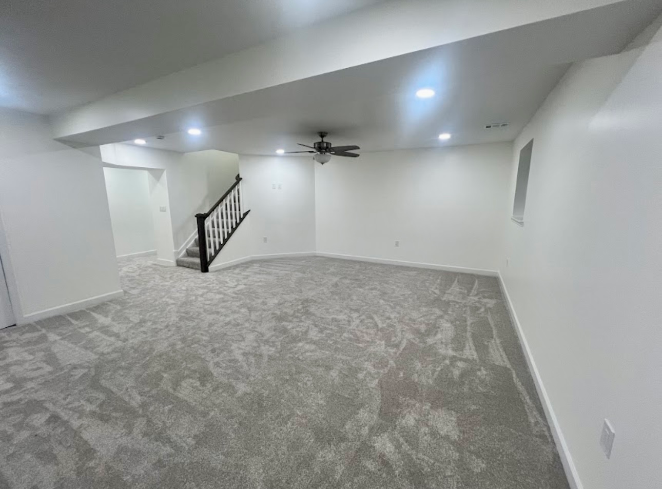 Empty carpeted living room with white walls, recessed lights, ceiling fan, and staircase railing.