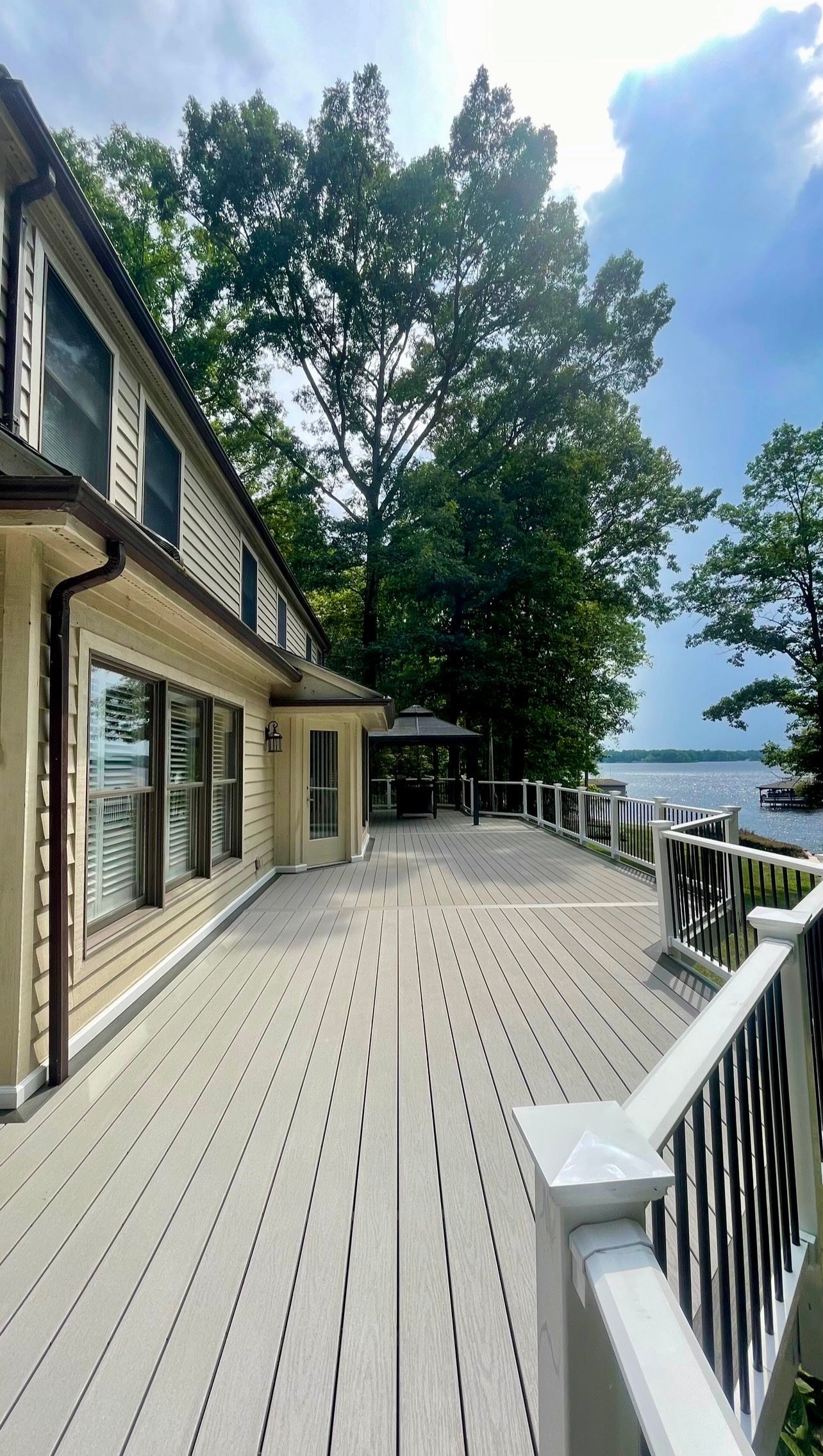 Wooden deck beside a house with white railing, overlooking trees and water under a blue sky.