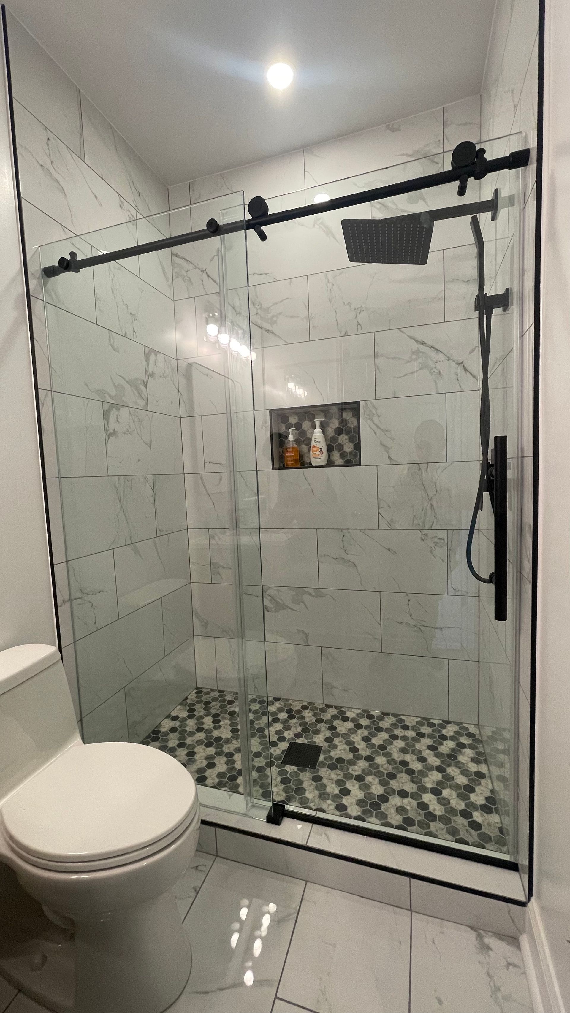 Modern bathroom with glass shower enclosure, white tiled walls, black hardware, and a patterned shower floor.
