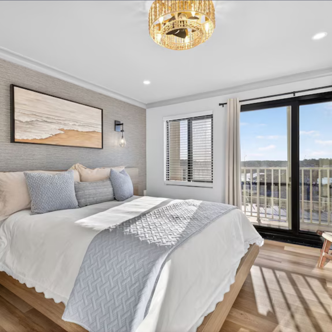 Bright bedroom with gray bedding, gold chandelier, and sliding glass doors to a balcony with city view