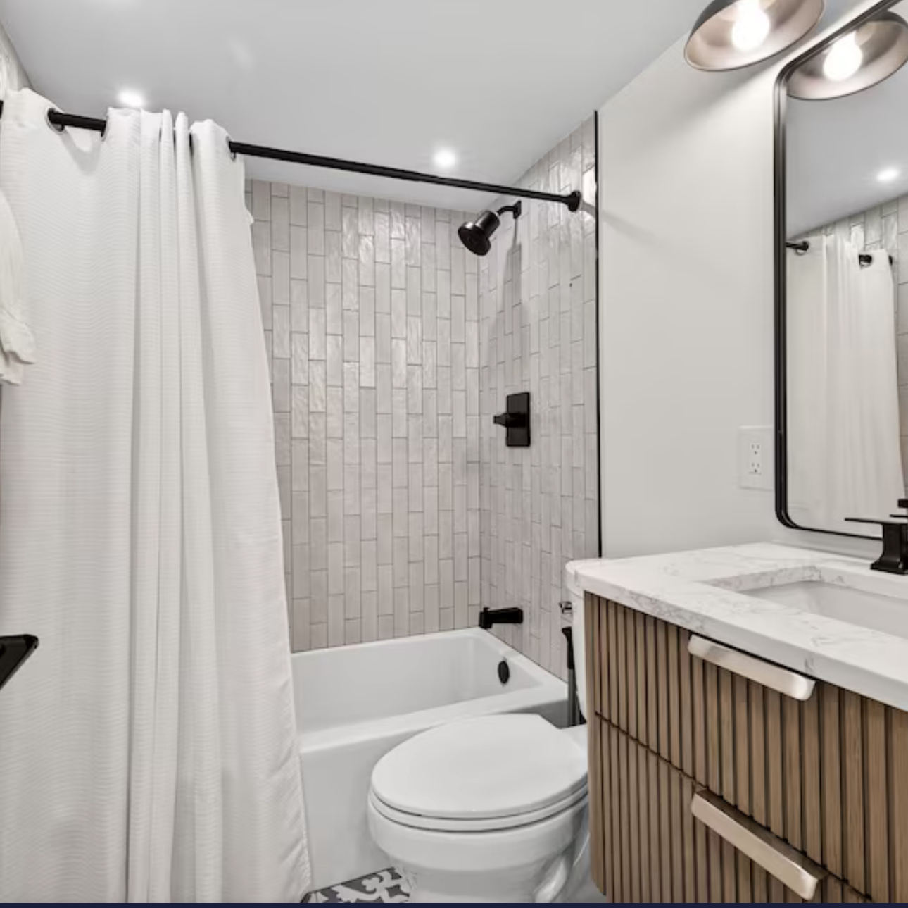 Bright white bathroom with tiled shower, tub, toilet, vanity, mirror, and black fixtures