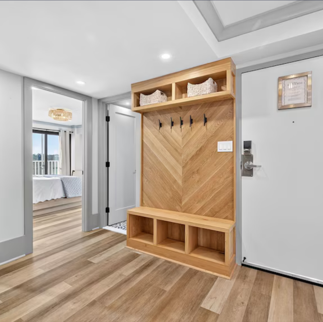 Entryway with built-in wooden coat rack and bench beside a white front door and hallway to a bedroom.