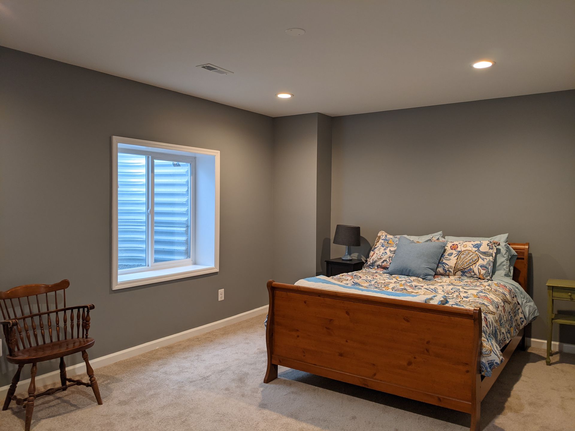 Sparse bedroom with gray walls, a wooden bed, a window, and a wicker chair near the corner.