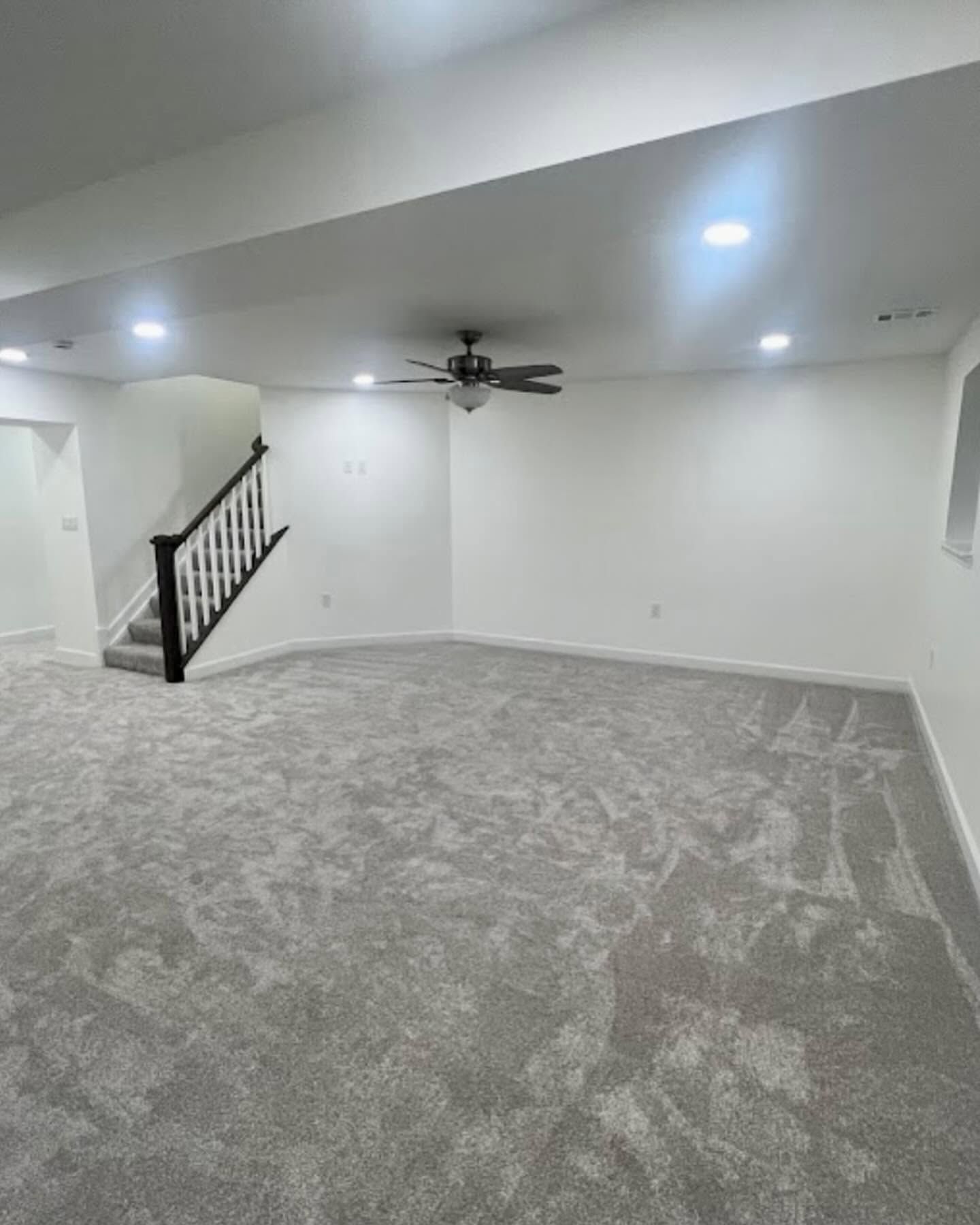 Empty carpeted living room with recessed lights, ceiling fan, and staircase railing in the corner.