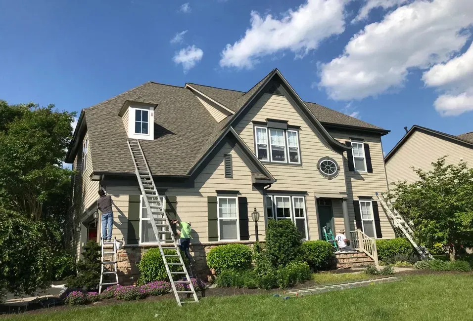 Two workers on ladders painting a tan two-story house with white trim and green shutters