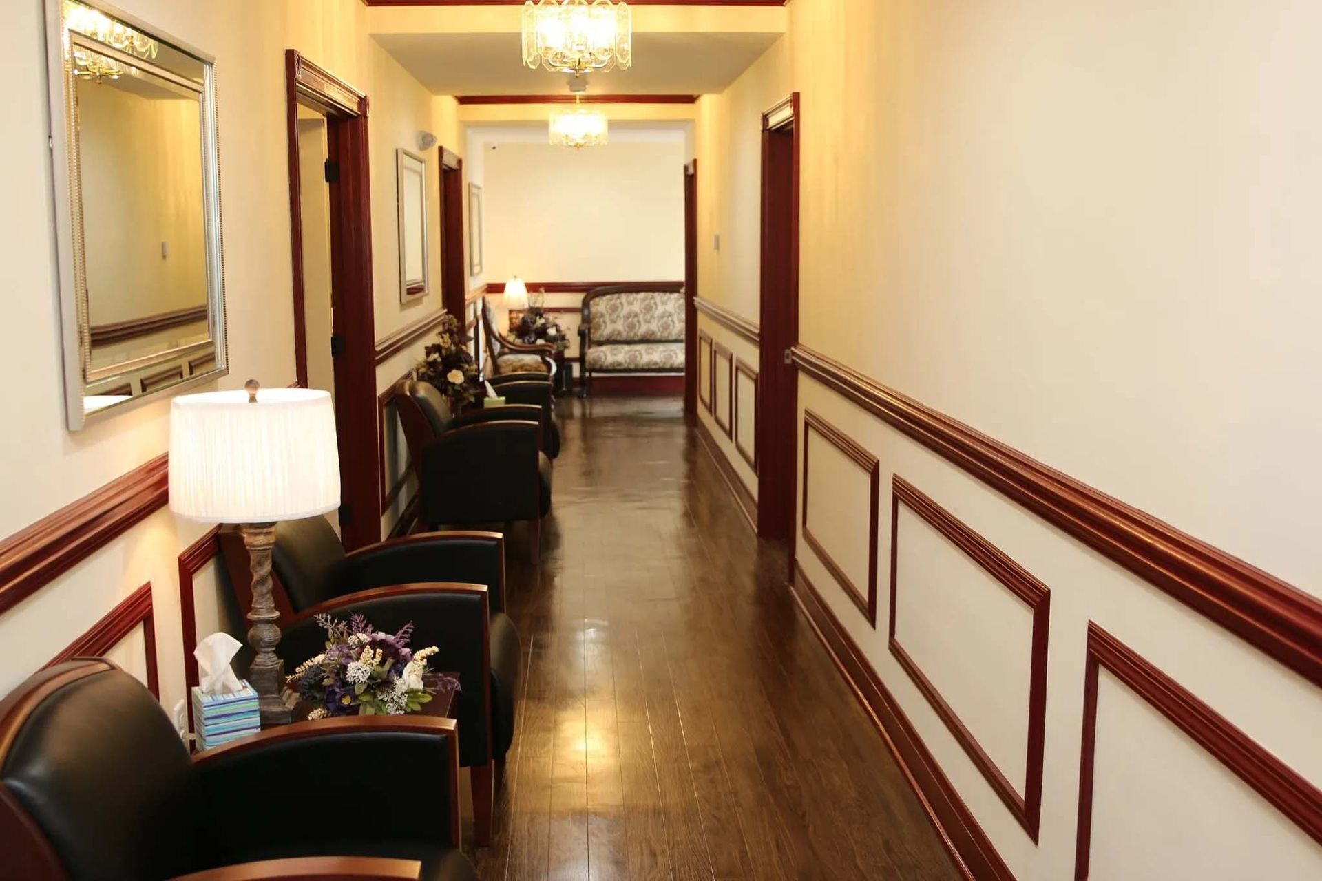 A long hallway with chairs , lamps and a mirror.