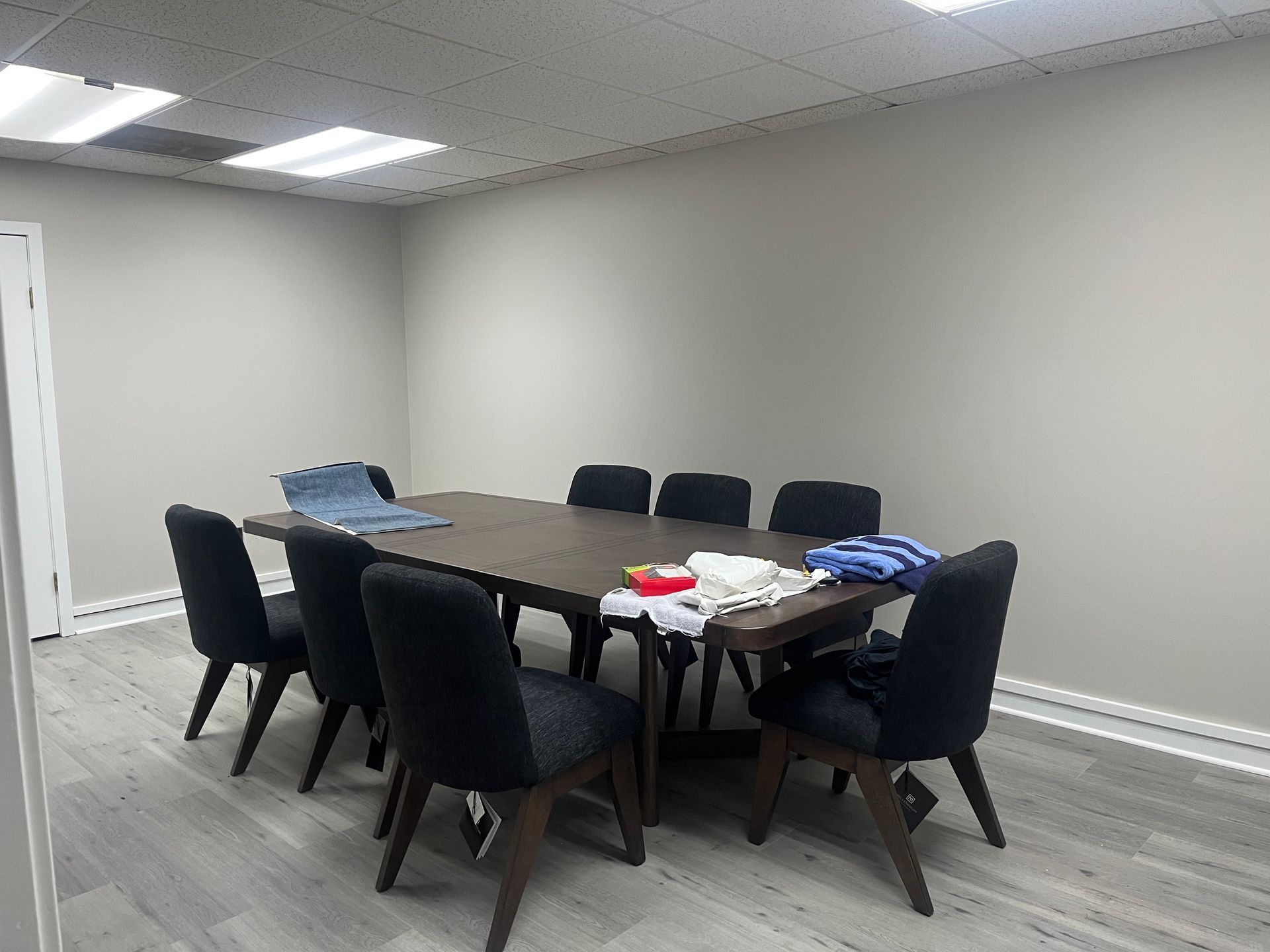 Empty conference room with a dark wood table, black chairs, and folded clothes on top, against beige walls.