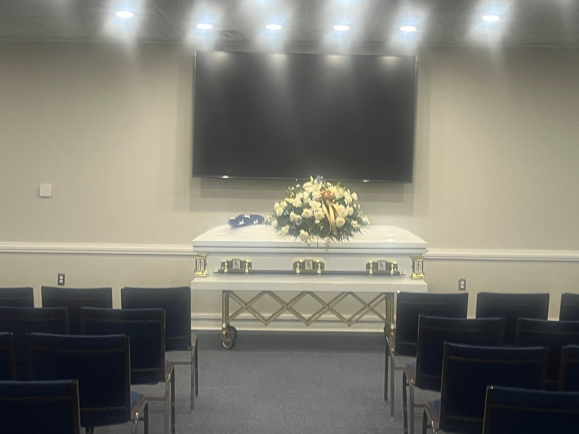 White casket with flowers in a chapel, viewed from rows of black chairs under soft lights