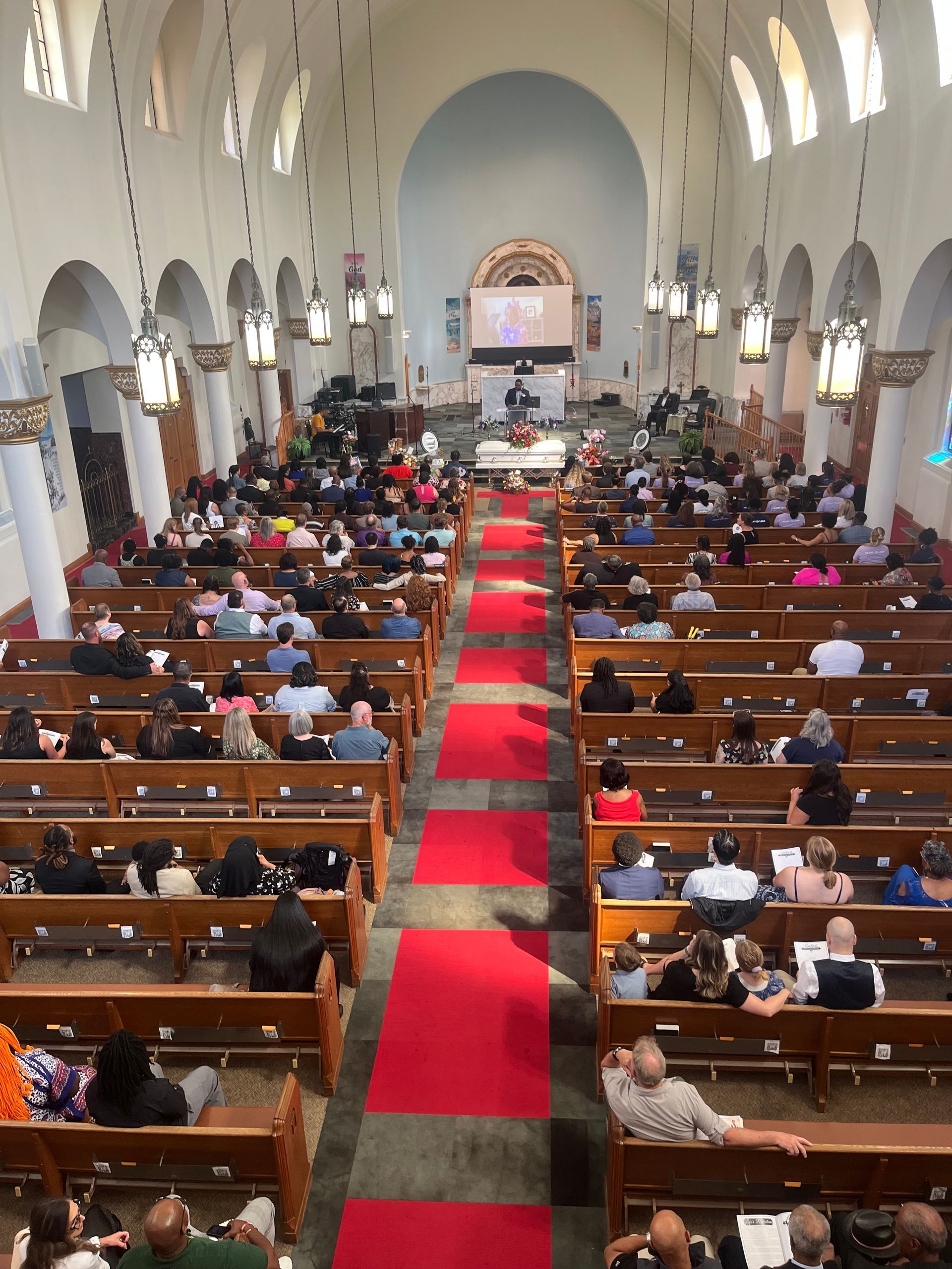 Crowded church interior with people seated in pews along a red carpet aisle leading to the altar