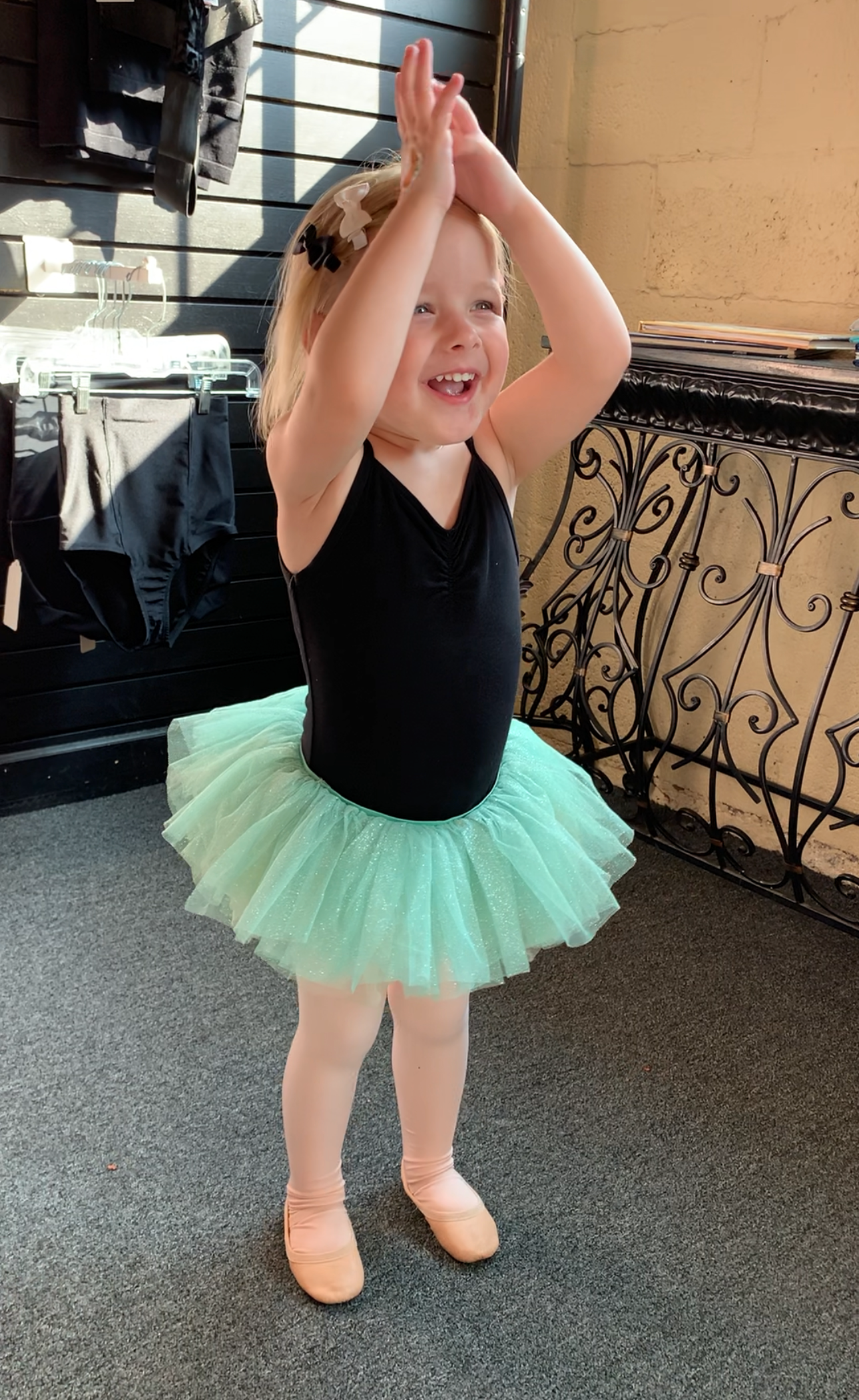Child in a black leotard and mint tutu clapping overhead, standing on a patio.