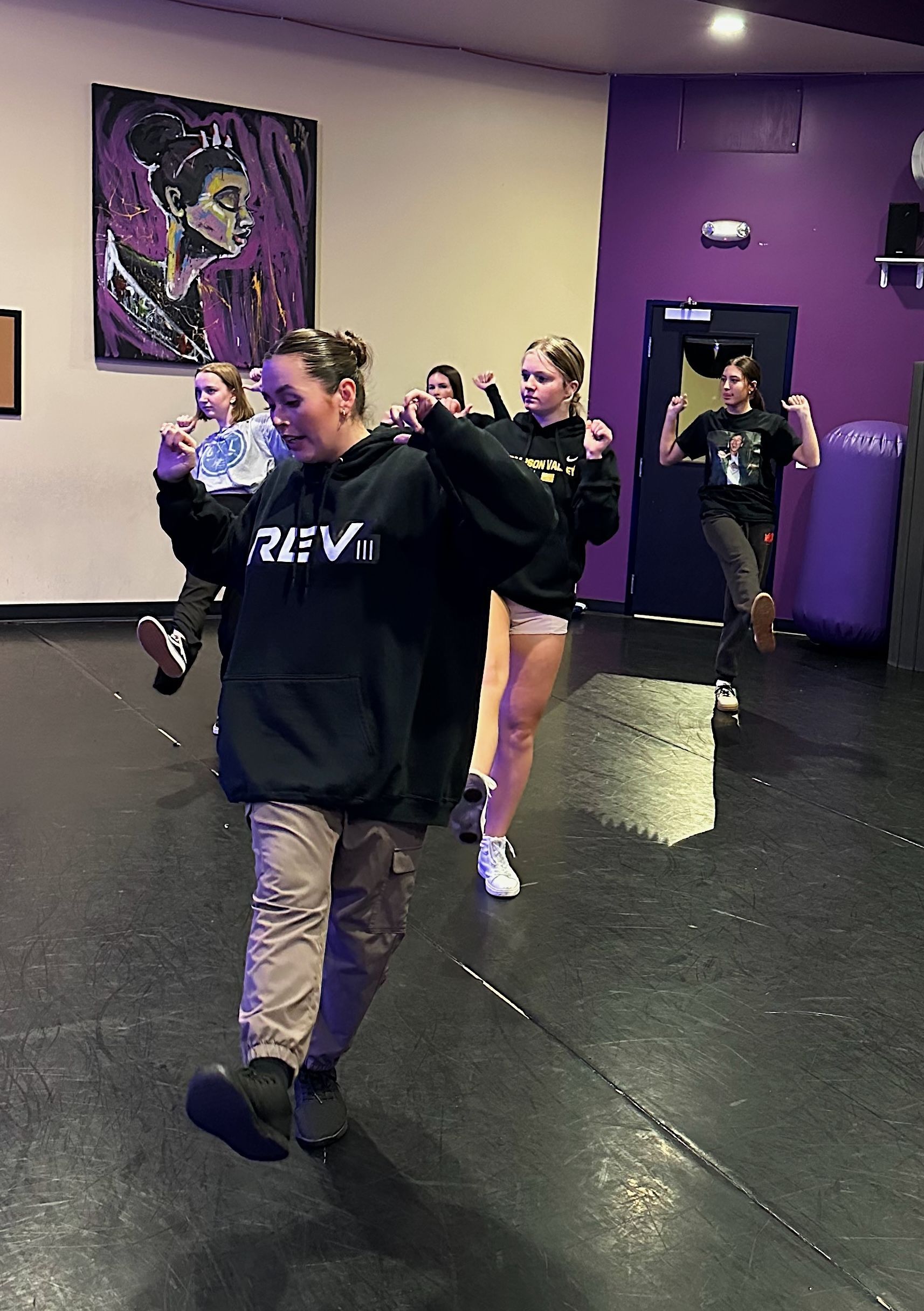 People practicing dance moves in a purple studio, led by someone in a black hoodie.