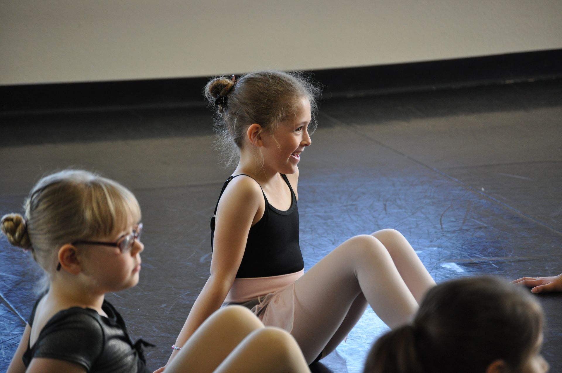 Children in dance class sitting on the floor, wearing black leotards and glasses, facing a mirrored wall.
