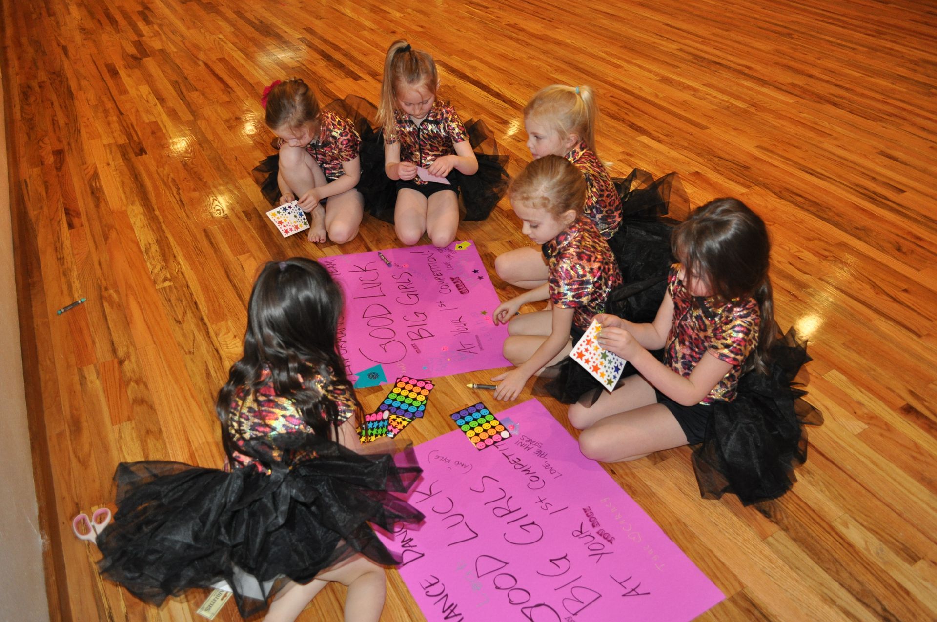 Children sitting in a circle on a wooden floor, writing on pink paper with colorful markers.