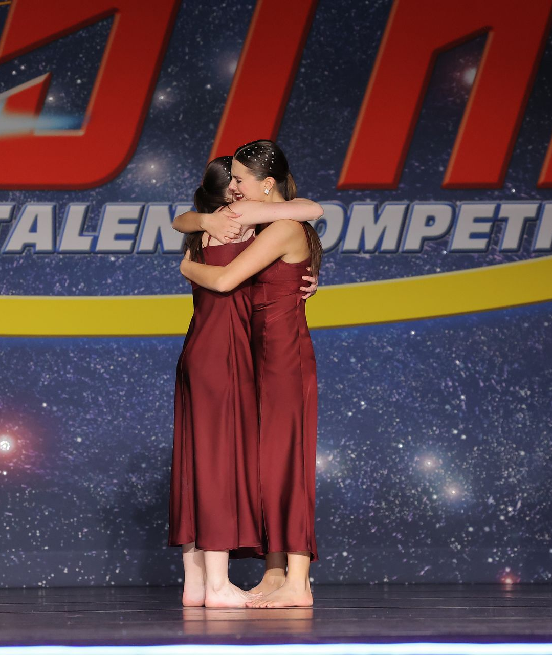 Two dancers in matching maroon costumes embrace on a competition stage