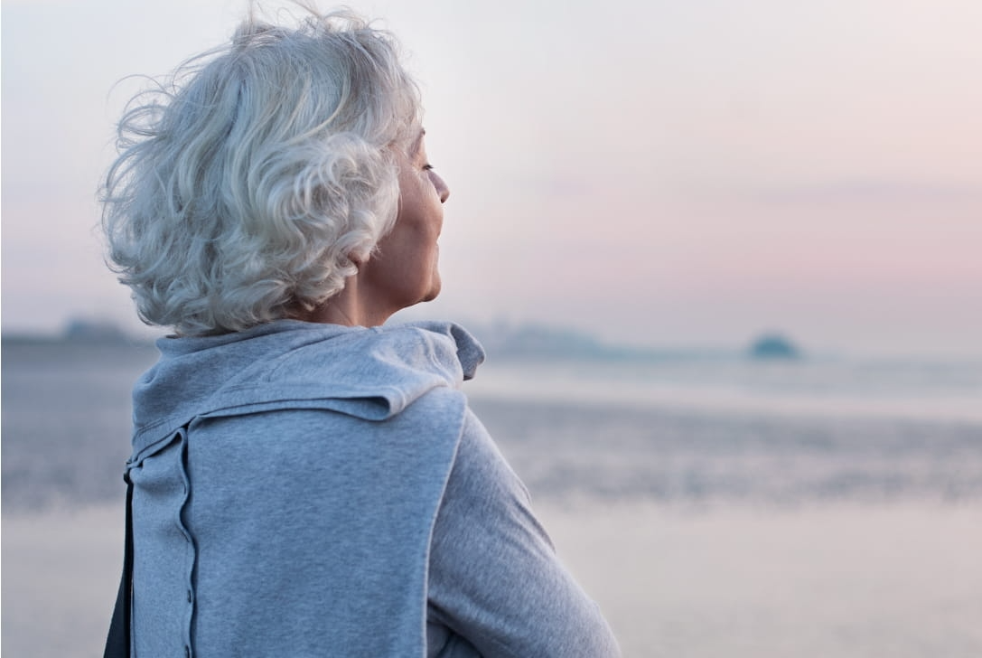Woman with white hair gazes out at a blurred, hazy body of water under a muted sky.