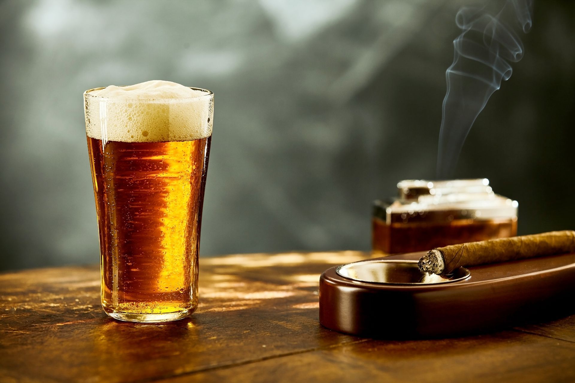 A foamy beer beside a lit cigar on a wooden table in warm, smoky light