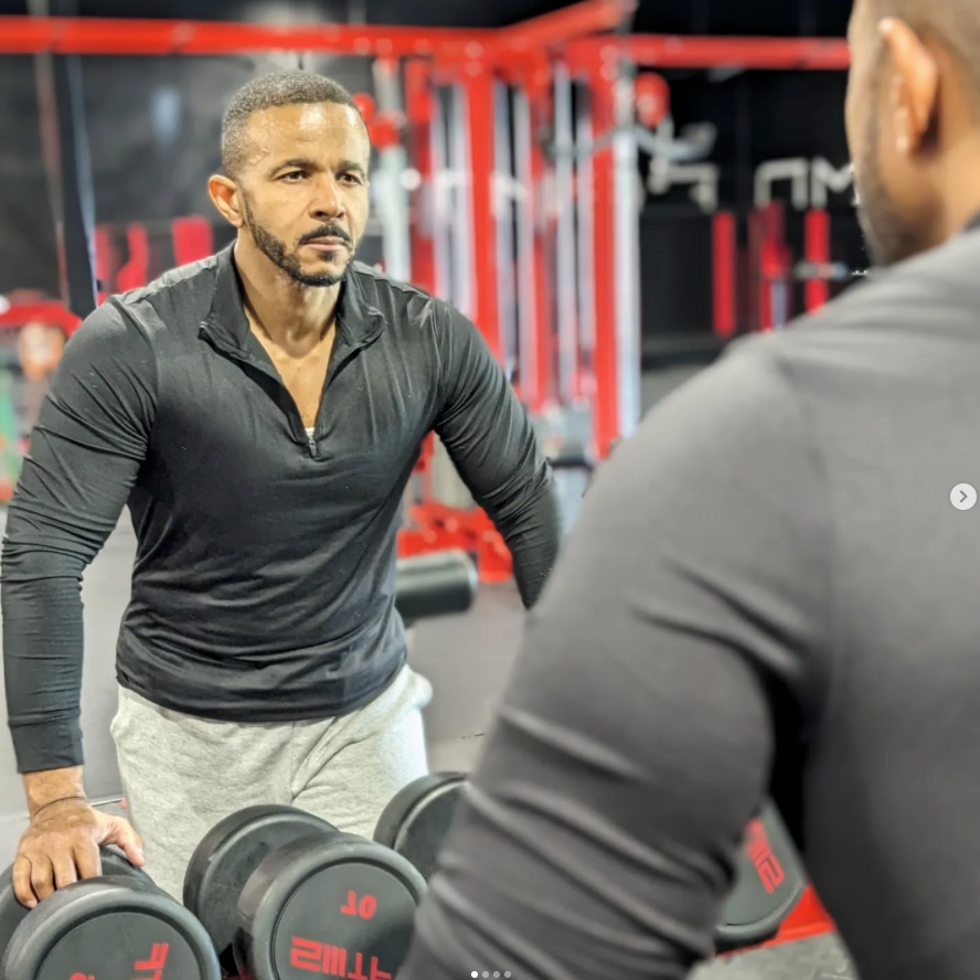 Two men in a gym, one leaning on dumbbells while facing a mirror.