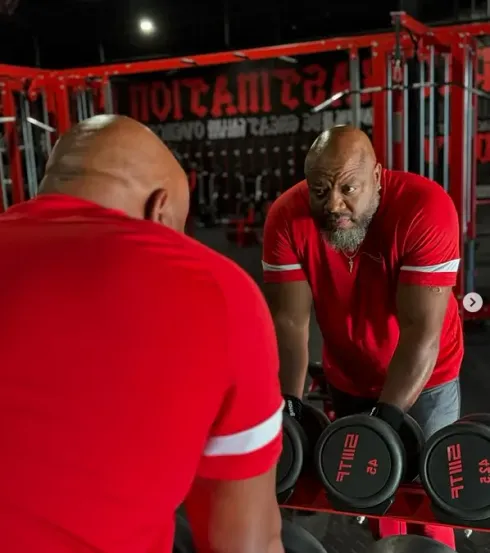 Man in a red shirt lifting dumbbells in a gym mirror, shown from behind and reflected in front.