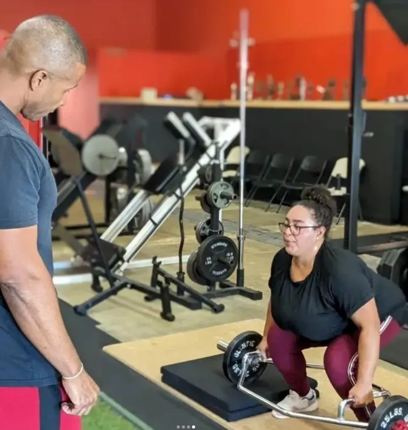 Gym trainer coaching a woman deadlifting on a platform, with exercise equipment in the background