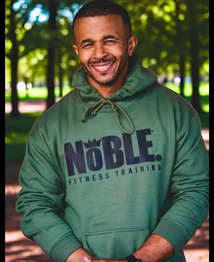 Man wearing a green hoodie with “NOBLE Fitness Training,” smiling outdoors in a park