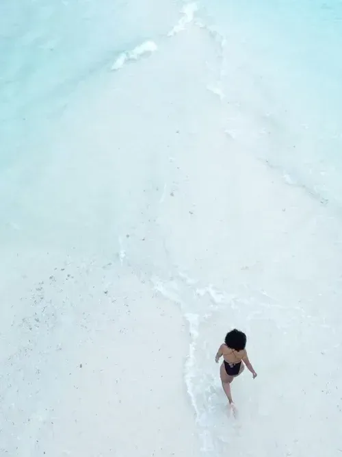 Person standing in shallow white surf on a pale turquoise beach, viewed from above