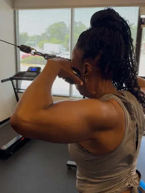 Woman flexing arm in a gym, with workout equipment and windows in the background
