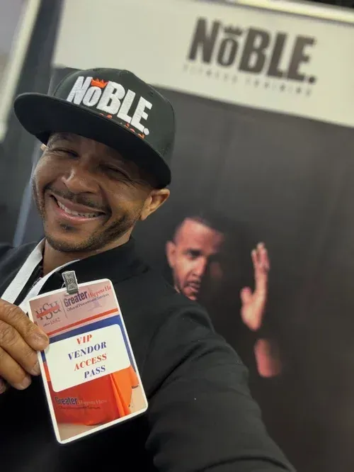 Smiling man in a black cap holds up a book and wears a “NOBLE” lanyard in a bookstore setting