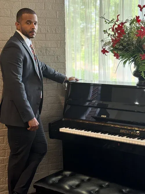 Man in a dark suit standing beside a black piano near a window with a vase of red flowers.