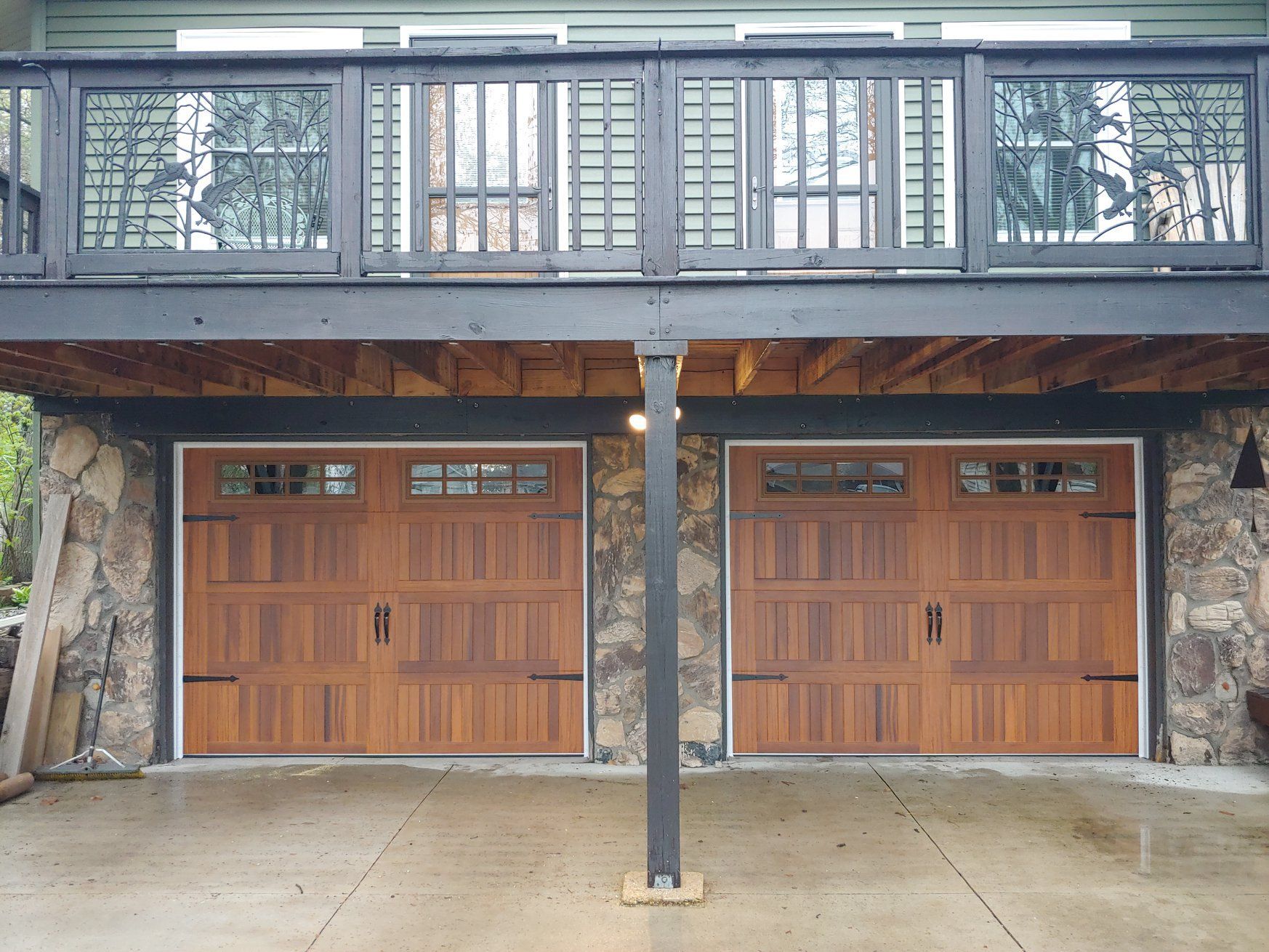 Two wooden garage doors beneath a balcony on a stone-faced house exterior.