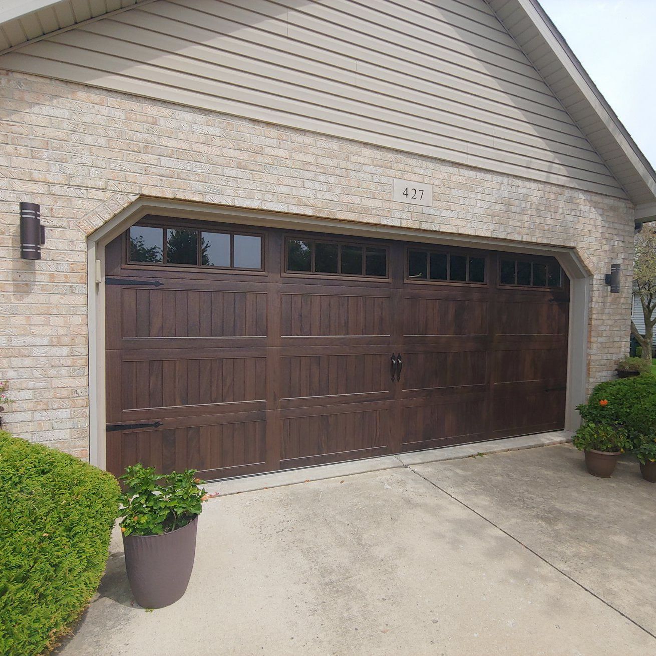 Brown paneled garage door on a beige brick house with a concrete driveway and potted plants
