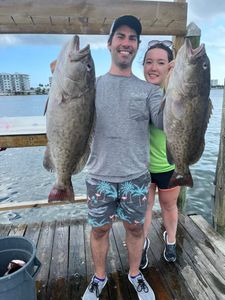Smiling couple on a dock holding two large fish, with water and buildings in the background.