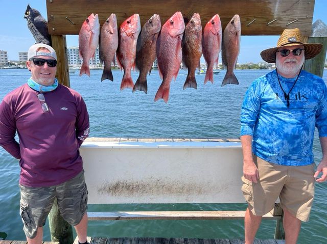 Two men on a boat deck with six red fish hanging between them, ocean in the background