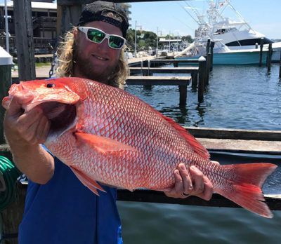 Person holding a large red fish at a marina, wearing sunglasses and a cap, with boats in the background