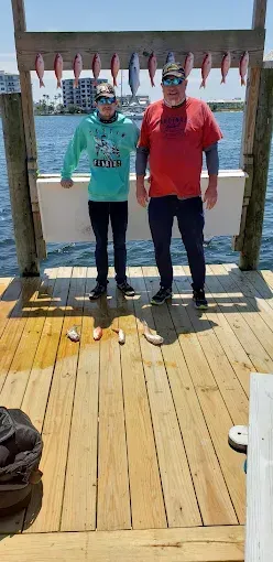 Two people standing on a wooden dock in the sun, with water and hanging fish behind them.