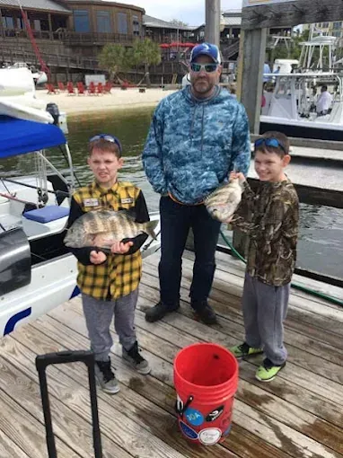 Three people on a dock holding large fish, with a red bucket in the foreground.