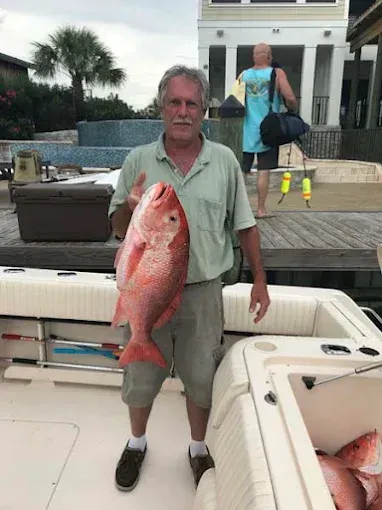 Man on a boat deck holding a large red fish, with waterfront buildings in the background