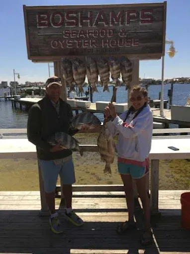 Two people holding fish at Boshamps Seafood & Oyster House dockside sign, with water and boats behind them