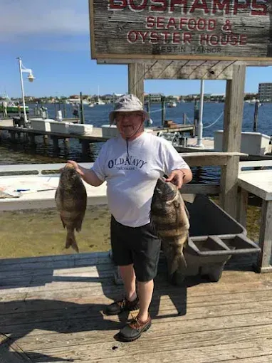 Person holding two large fish on a dock by the water with a seafood sign overhead