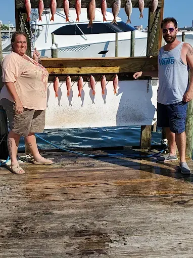 Two people posing beside a docked fish cleaning station with many fish hanging to dry in the sun
