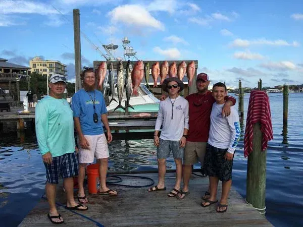Five people posing on a dock with two large fish hanging between them by the water under a blue sky.