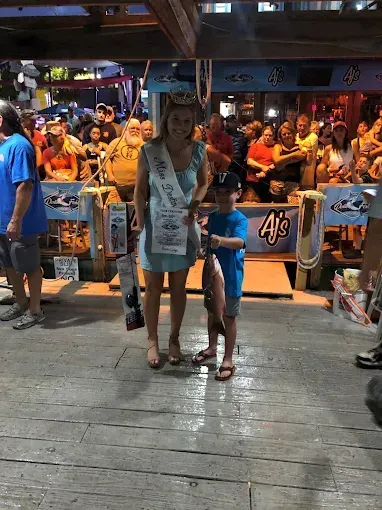 Two people standing on a wooden boardwalk in front of a crowded, brightly lit bar.