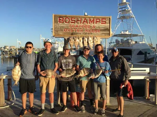 Six people holding fish pose on a dock in front of a Boshamps seafood sign and boat.