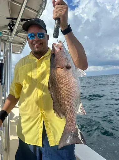 Person on a boat holding up a large fish, wearing a yellow shirt and sunglasses, with open water behind.