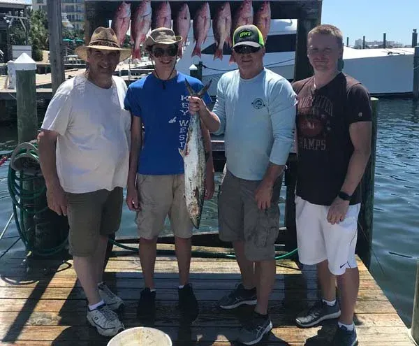 Four people stand on a dock by the water, posing in front of hanging fish.