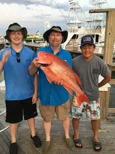 Three people on a dock hold a large red fish, smiling near boats at sunset.