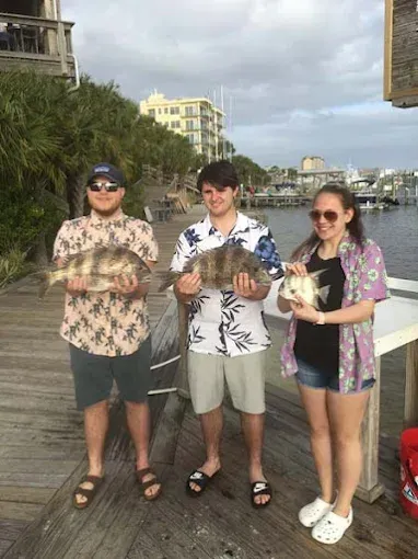 Three people holding fish on a seaside road, smiling in casual summer clothes.