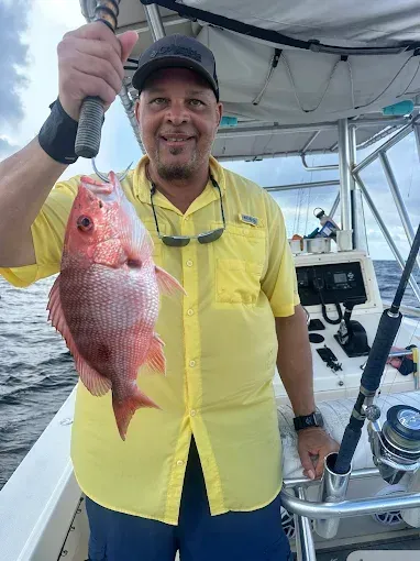 Angler on a boat holding a large red fish, wearing a bright yellow shirt and cap.