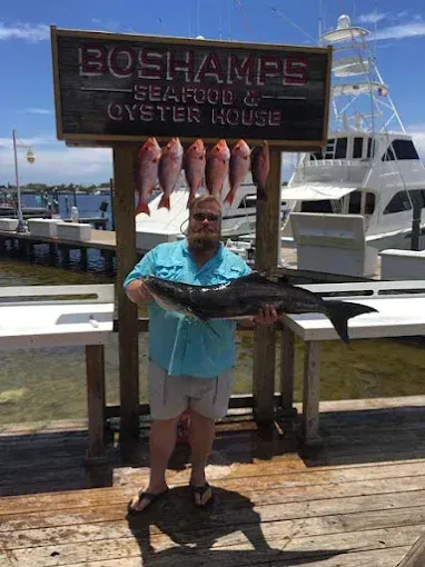 Man holding a large fish in front of a seafood restaurant sign on a dock by the water