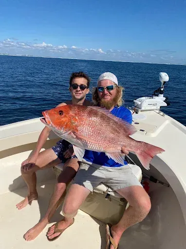 Two people on a boat holding a large red fish, with blue ocean and sky in the background.